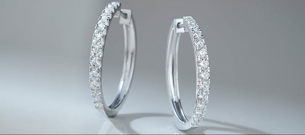 Pair of silver hoop earrings with clear gemstones on a reflective surface.